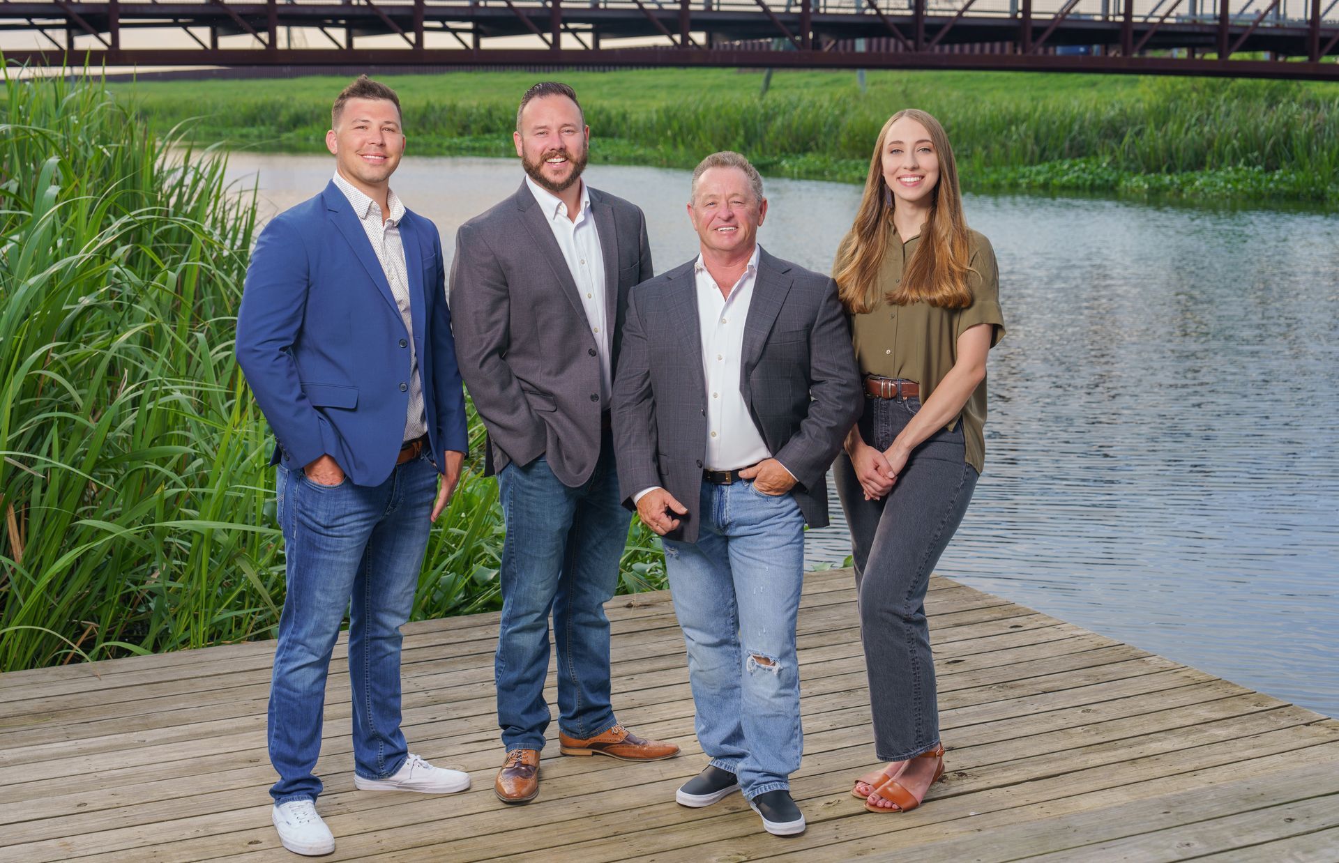 Four people pose on a wooden dock by water. Three men in blazers and jeans, one woman in a shirt and pants.
