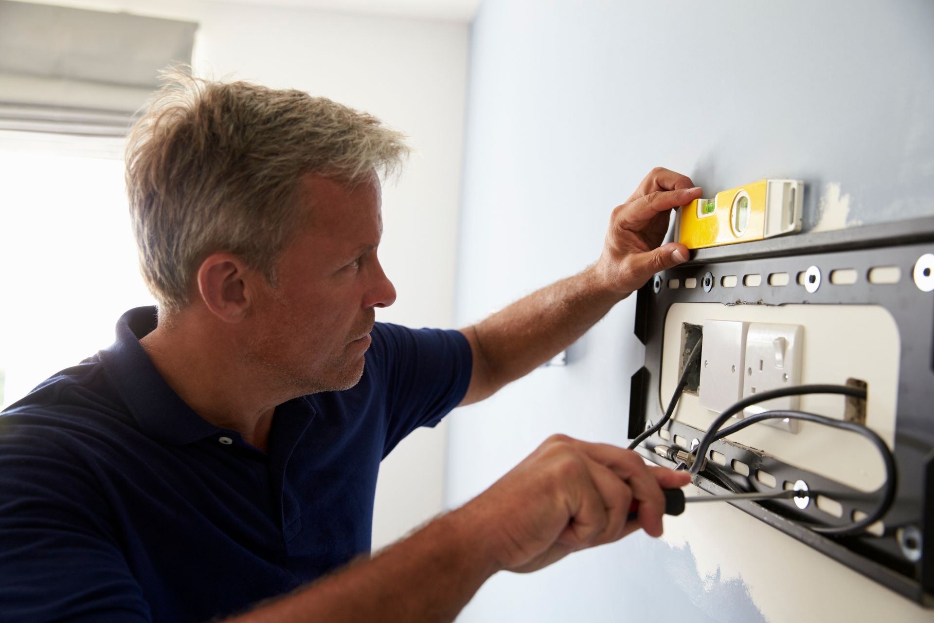 Man using level and screwdriver to install TV mount on blue wall.