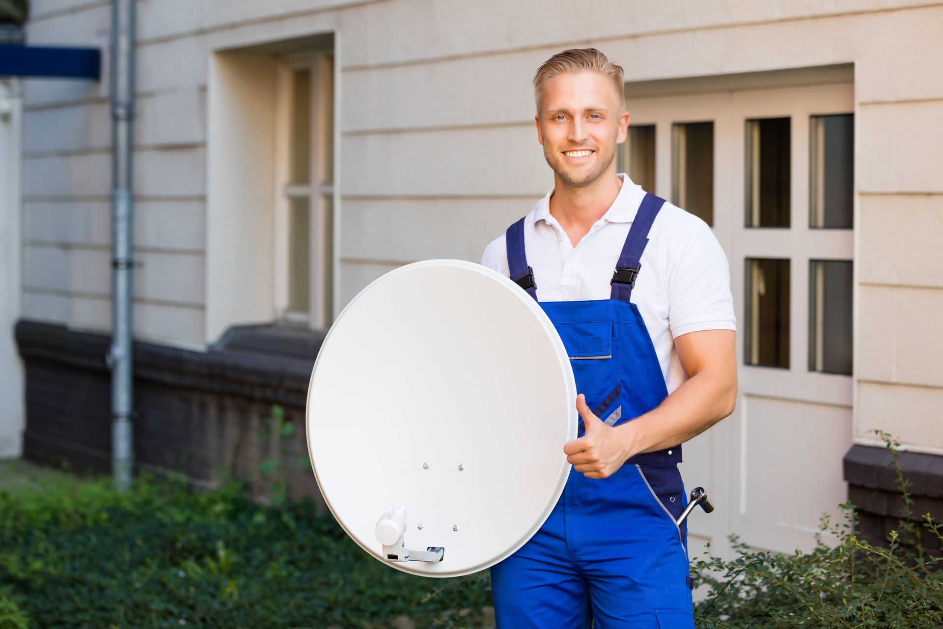 Man in blue overalls holding a satellite dish, smiling in front of a building.