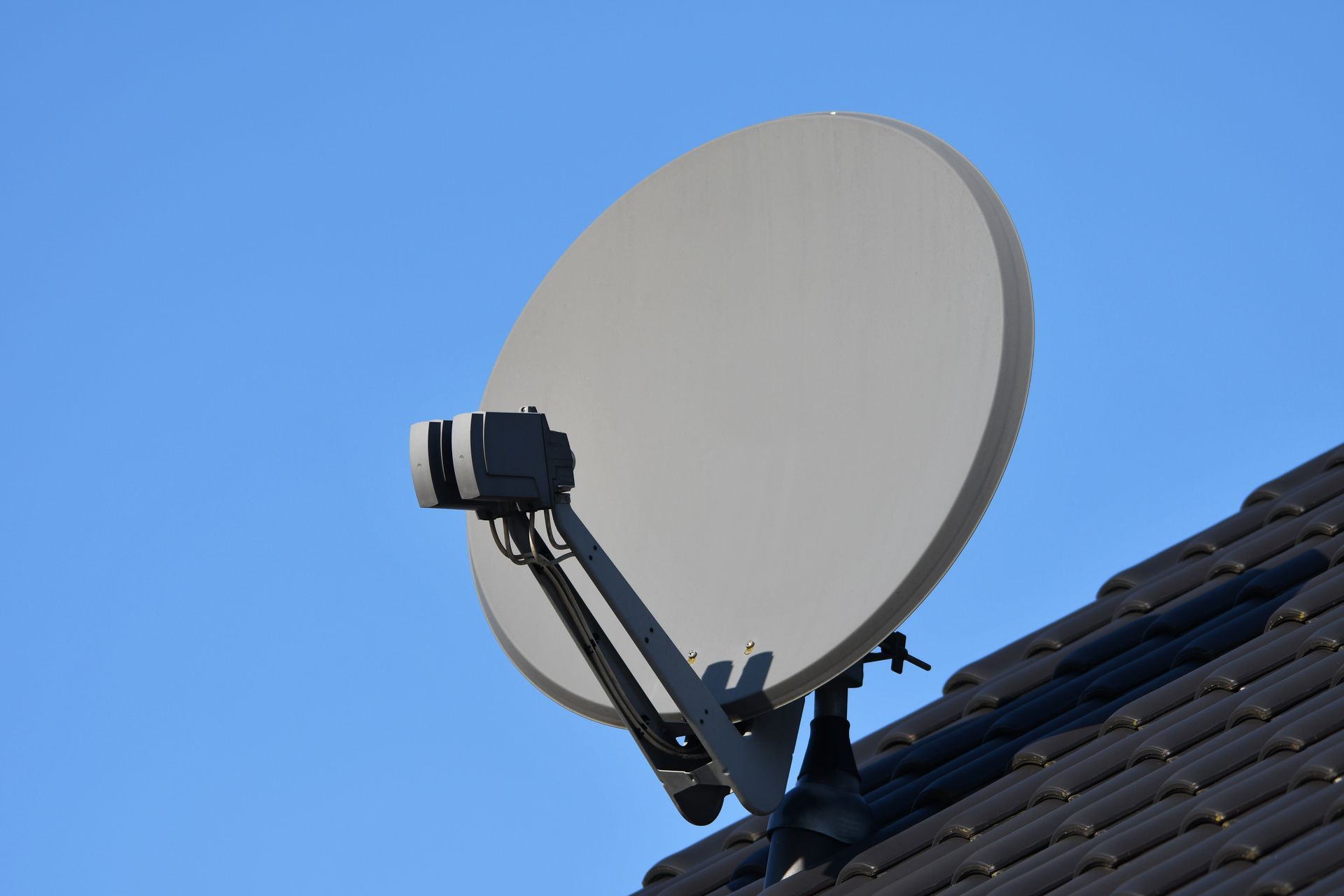 Satellite dish mounted on a dark tiled roof against a bright blue sky.