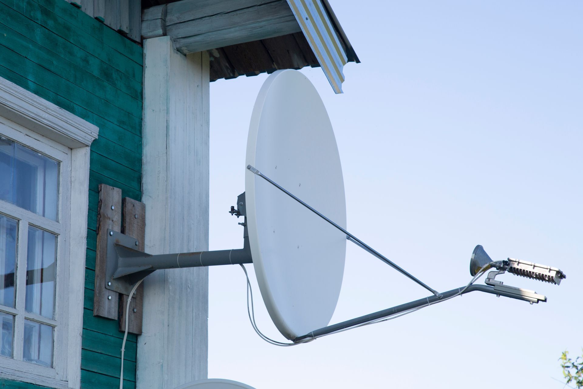 White satellite dish attached to the green wooden side of a house.