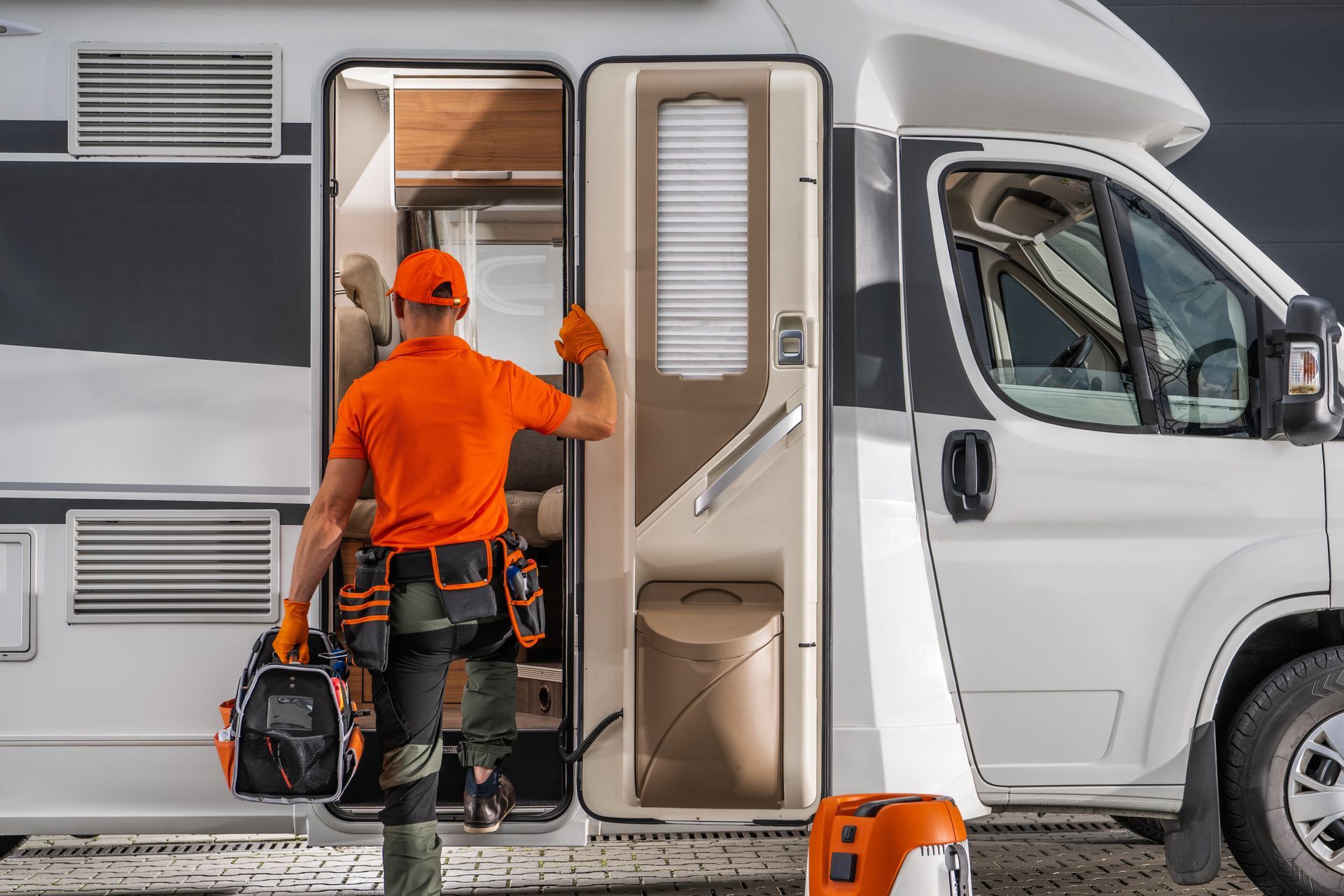 Person in orange shirt and tool belt enters a white RV, holding a toolbox.