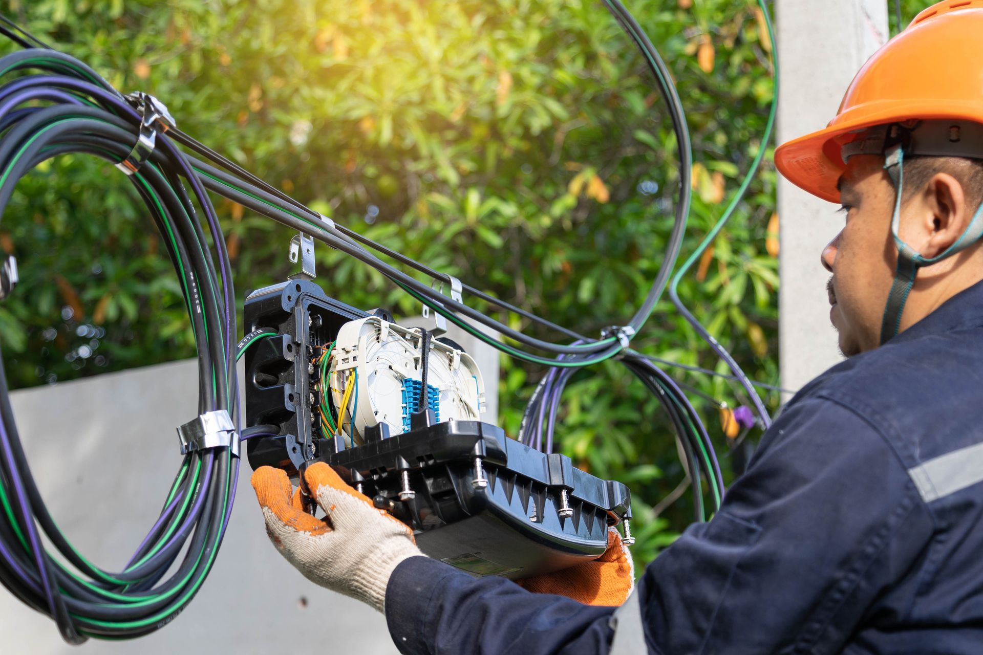 Worker in orange hard hat connecting fiber optic cables on utility pole.