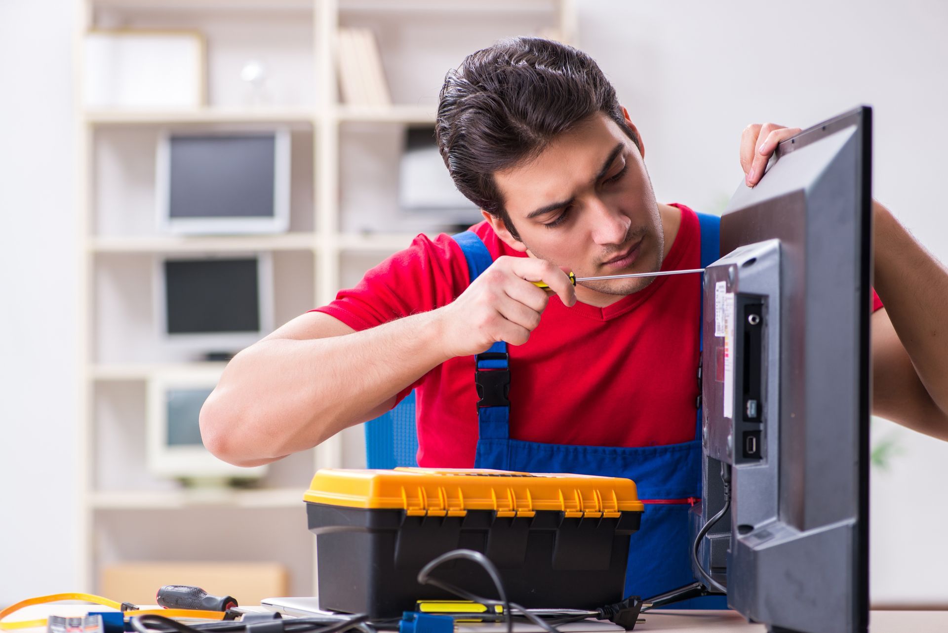 Man in red shirt and blue overalls fixing a TV with a screwdriver in a well-lit room.