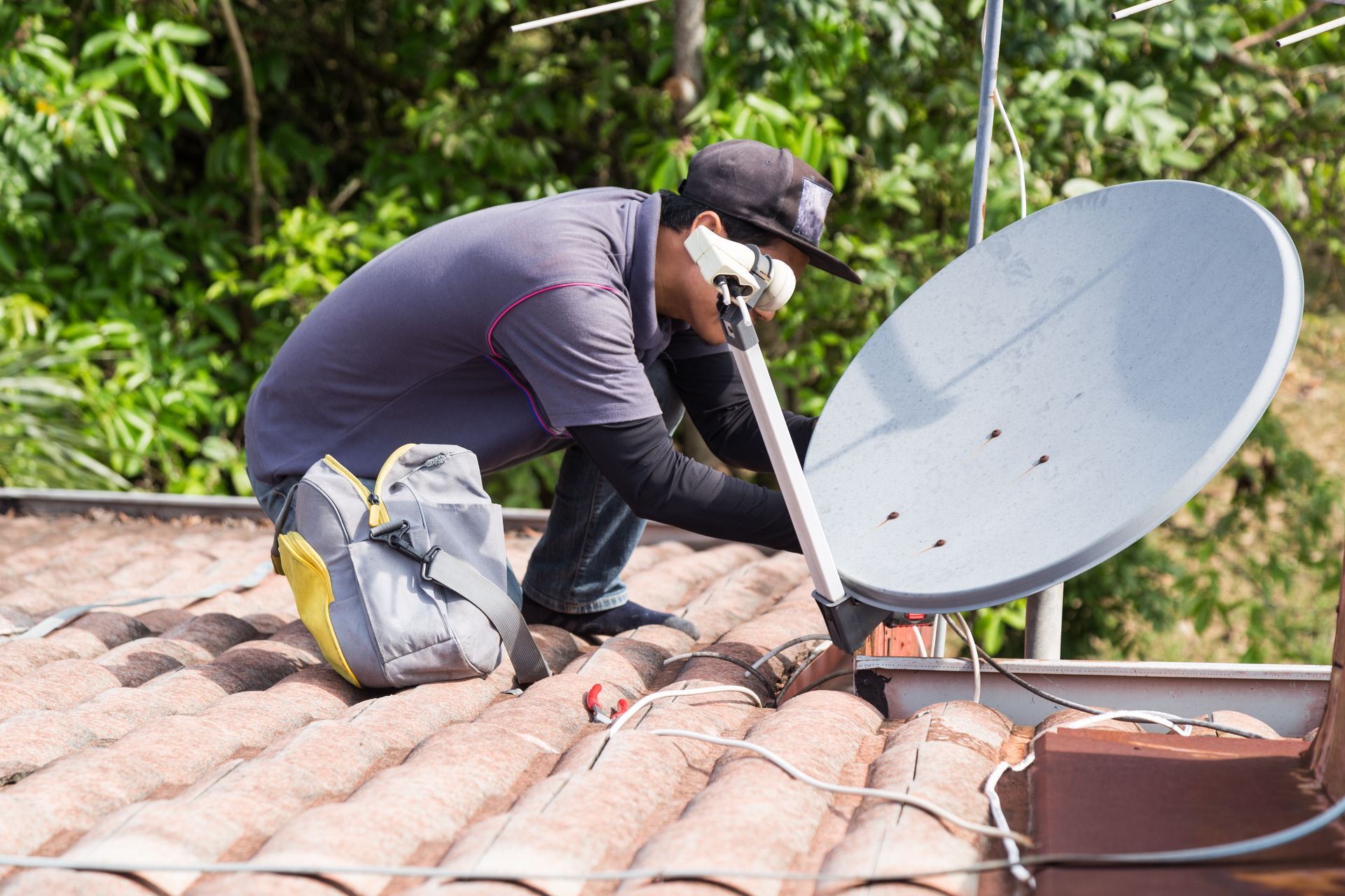 A person installing a satellite dish on a red-tiled roof, near trees.