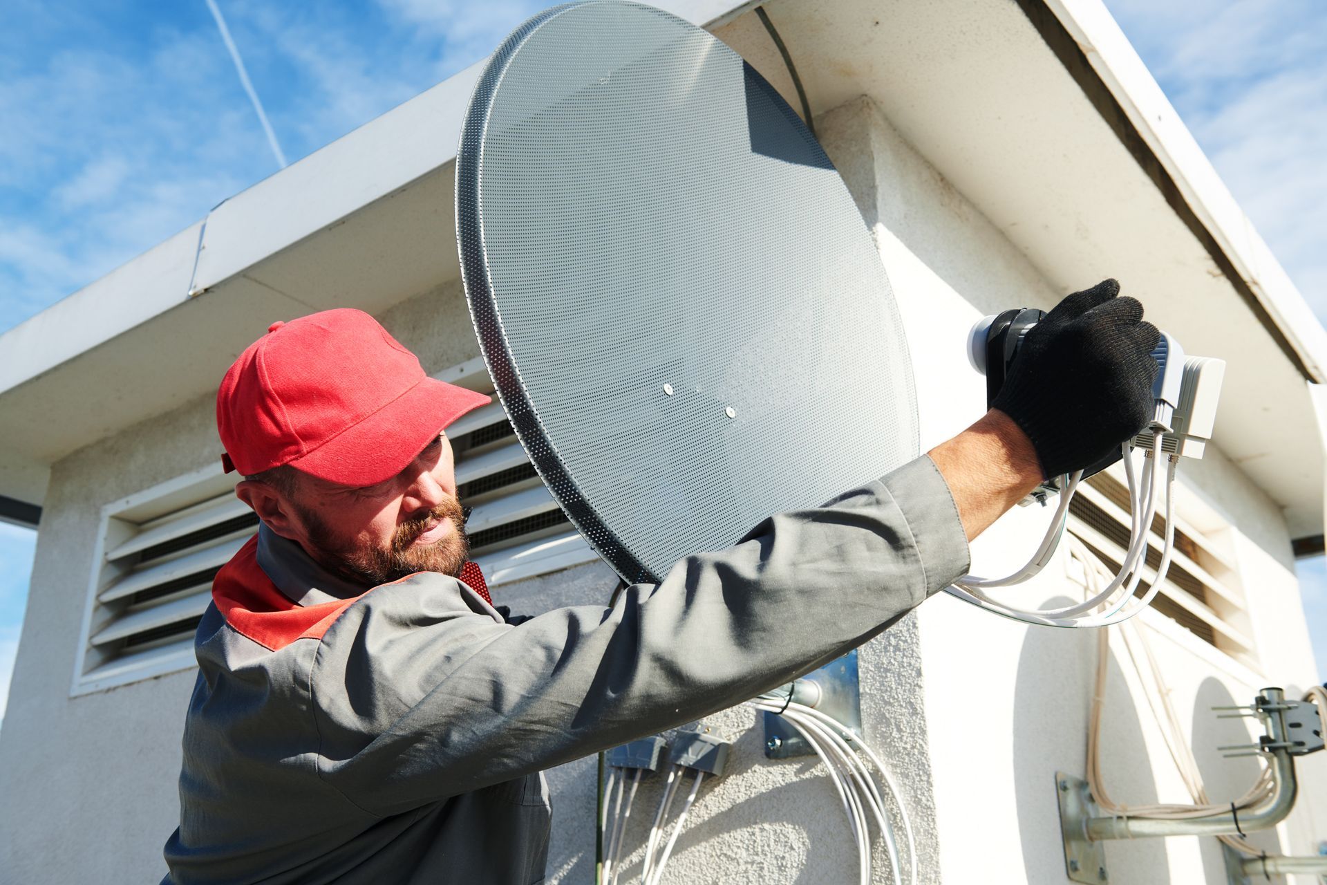 Man in red cap installs a satellite dish on a building exterior.