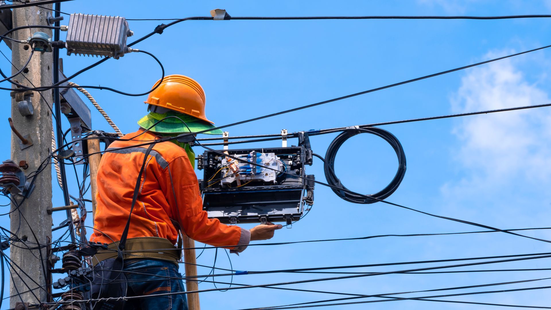 Lineman in orange suit working on utility pole with wires and cables against blue sky.