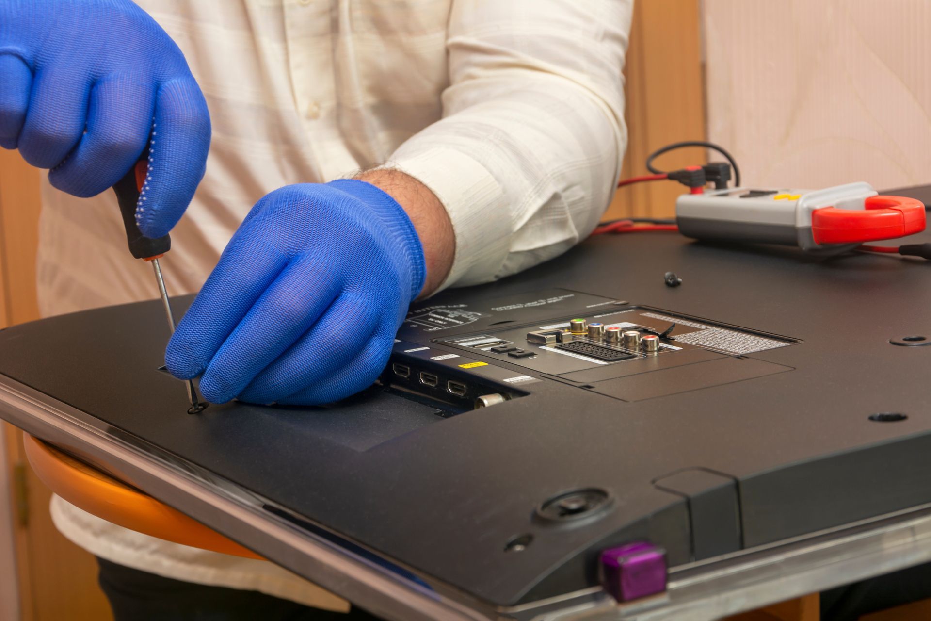 A person wearing blue gloves repairs a TV with a screwdriver, back panel open, in a room.