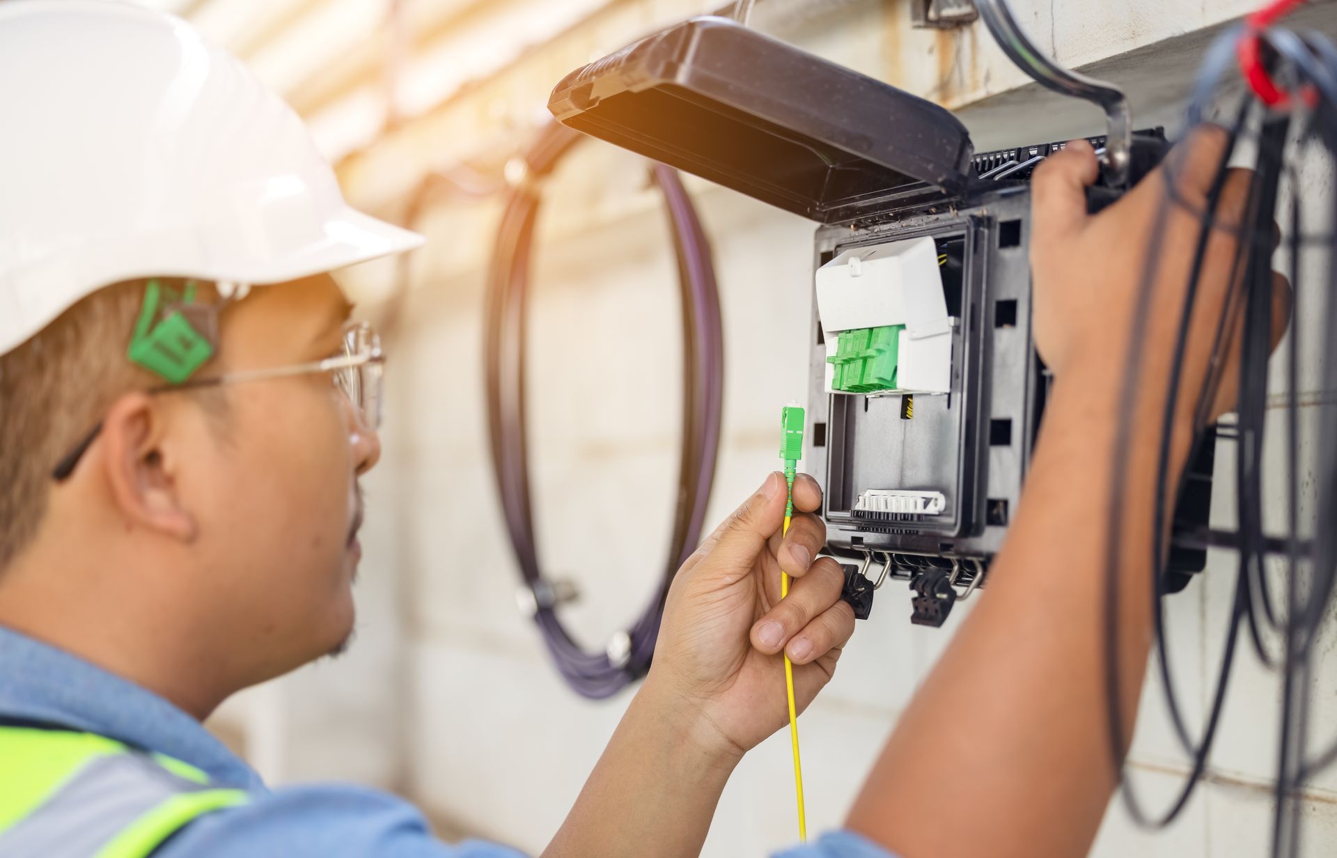 A technician in a hard hat connecting a fiber optic cable to a network box on a wall.