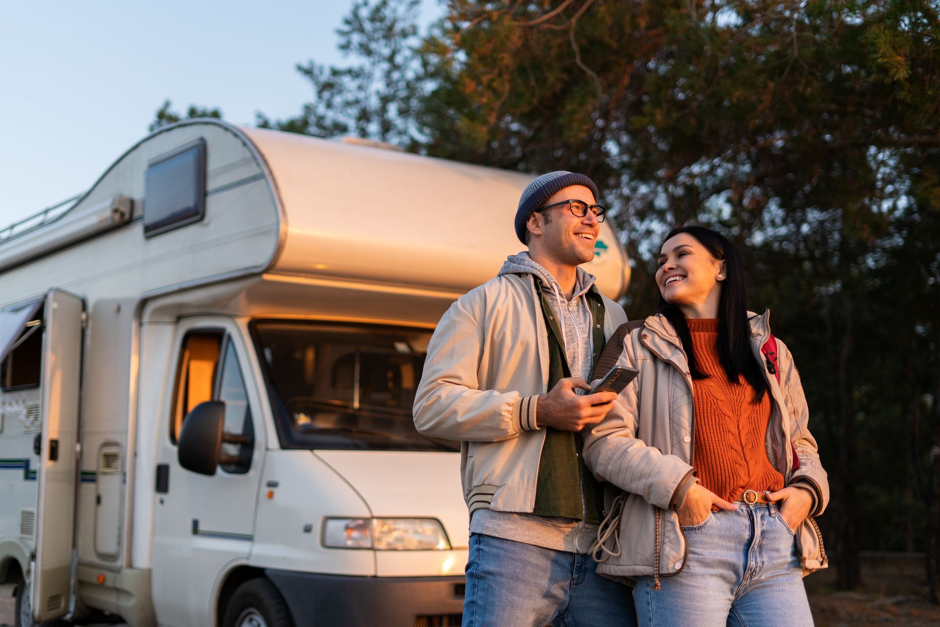 Couple standing near a white RV, smiling and looking upward in a natural setting.