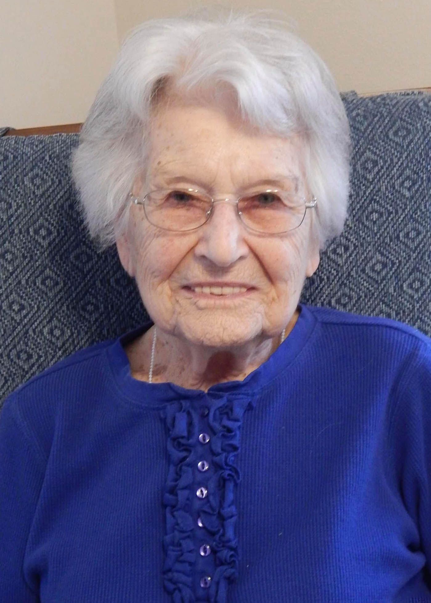 An elderly woman wearing glasses and a blue shirt is sitting in a chair.