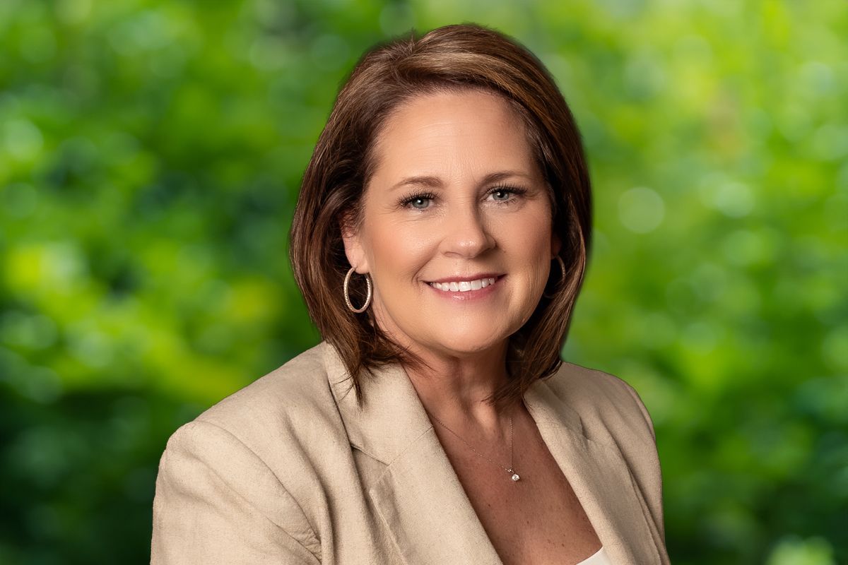 Woman in tan blazer smiles against a blurred green background.