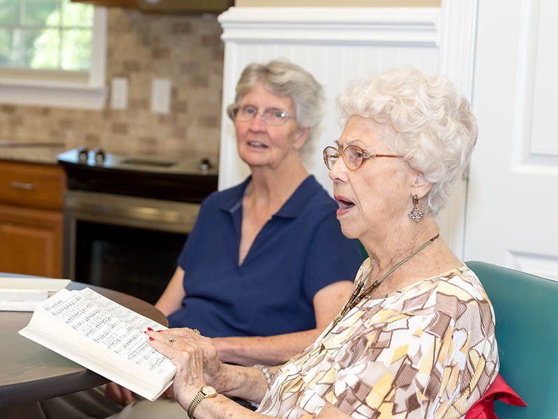 Two older women are sitting at a table reading a book.