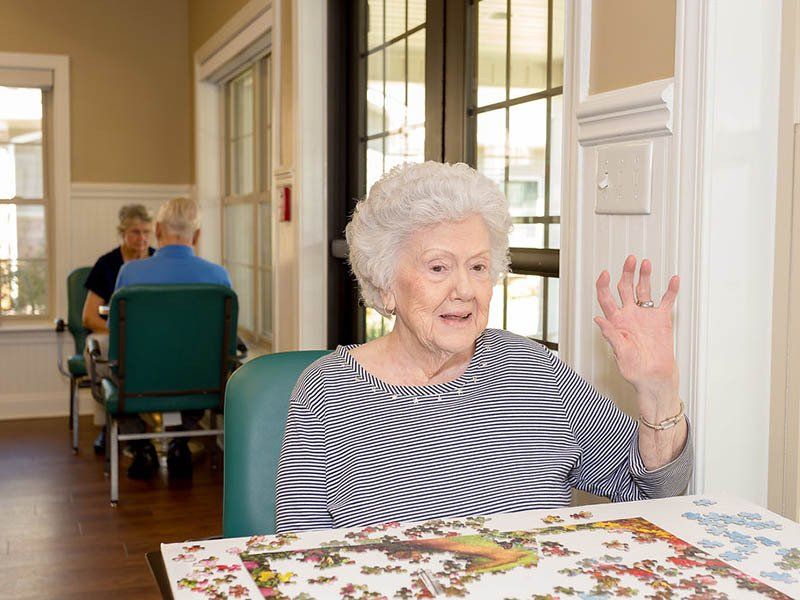 An elderly woman is sitting at a table with a puzzle and waving.