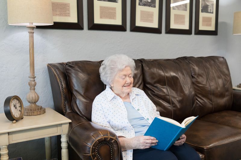 An elderly woman is sitting on a couch reading a book.