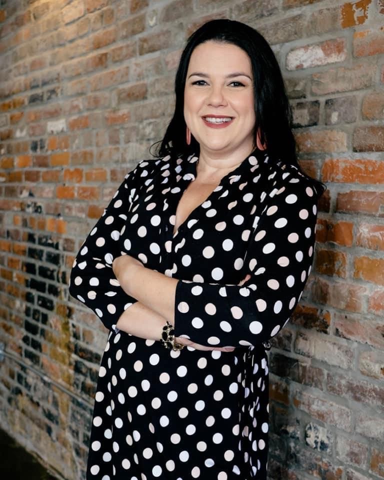 A woman in a black and white polka dot dress is standing in front of a brick wall.  life coach training, the coaching guild