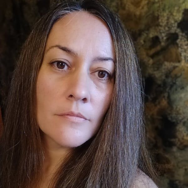 A woman with long hair is standing in front of a stone wall and looking at the camera. Life Coach Caroline Meyers, the coaching guild,  life coach training