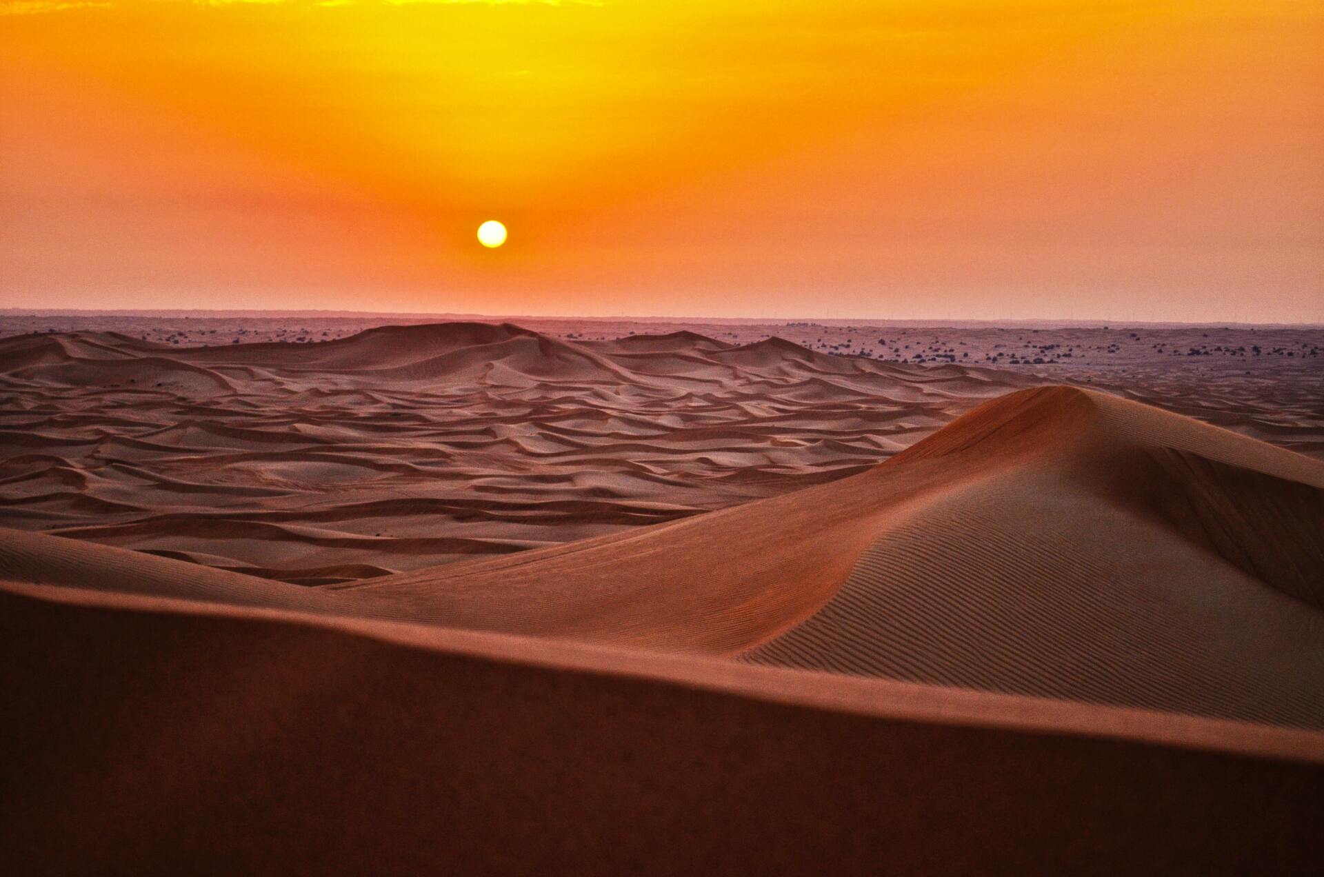 The sun is setting over a desert landscape with sand dunes in the foreground.