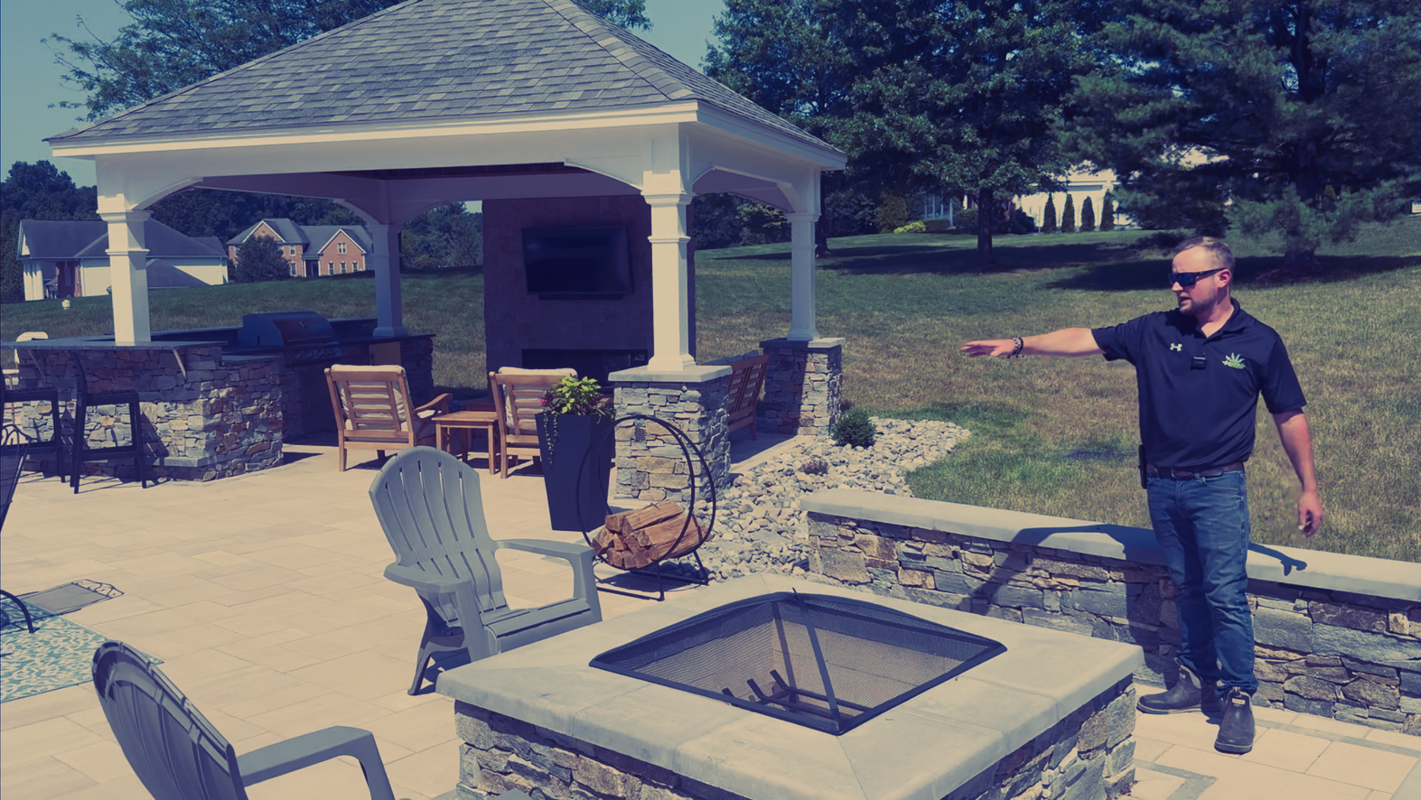 Man points toward a patio with fire pit, seating, and gazebo in a sunny outdoor setting.
