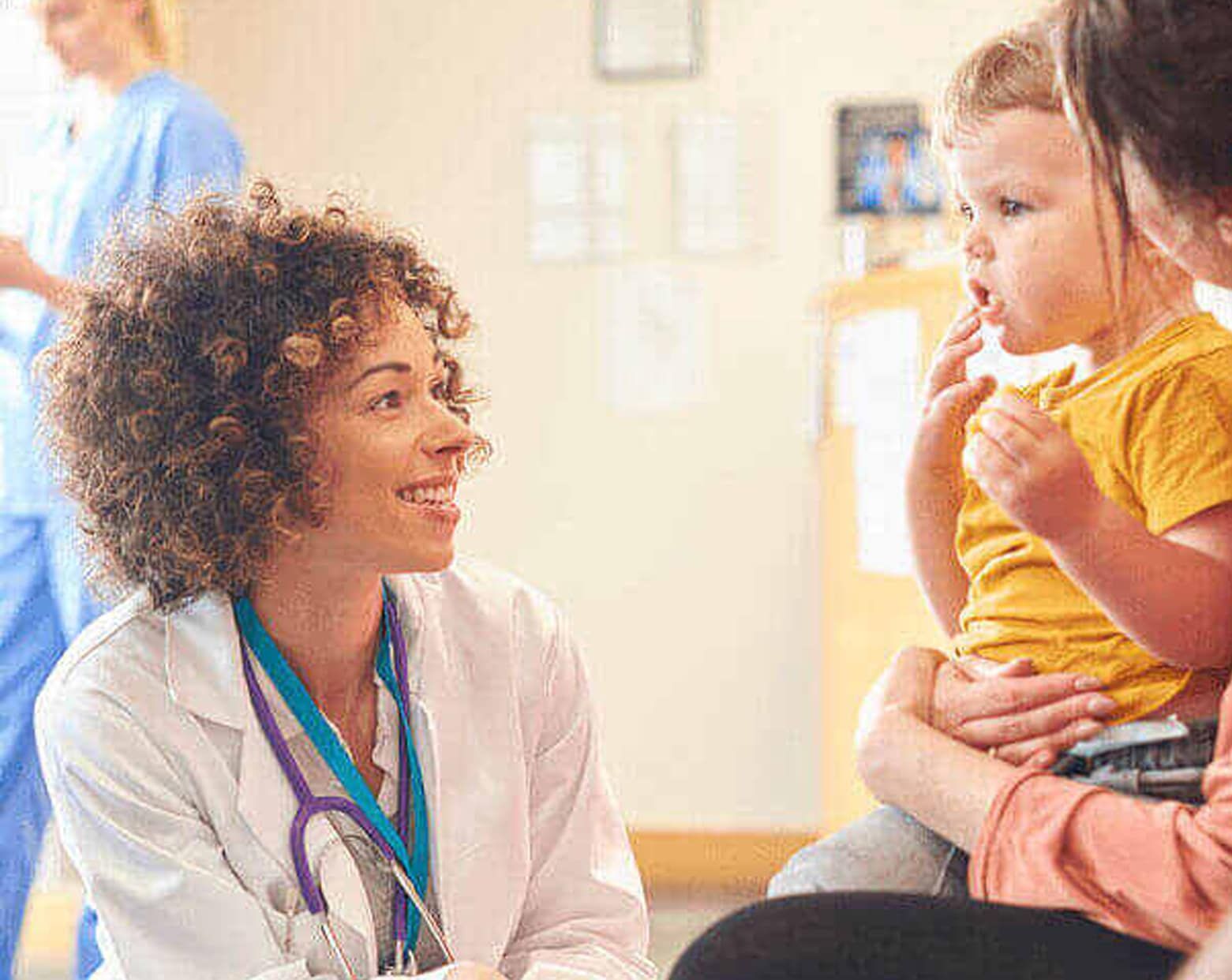 Doctor smiles at a toddler held by a woman in a medical office; a nurse stands in the background.