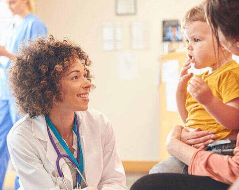 Doctor smiles at a toddler held by a woman in a medical office; a nurse stands in the background.