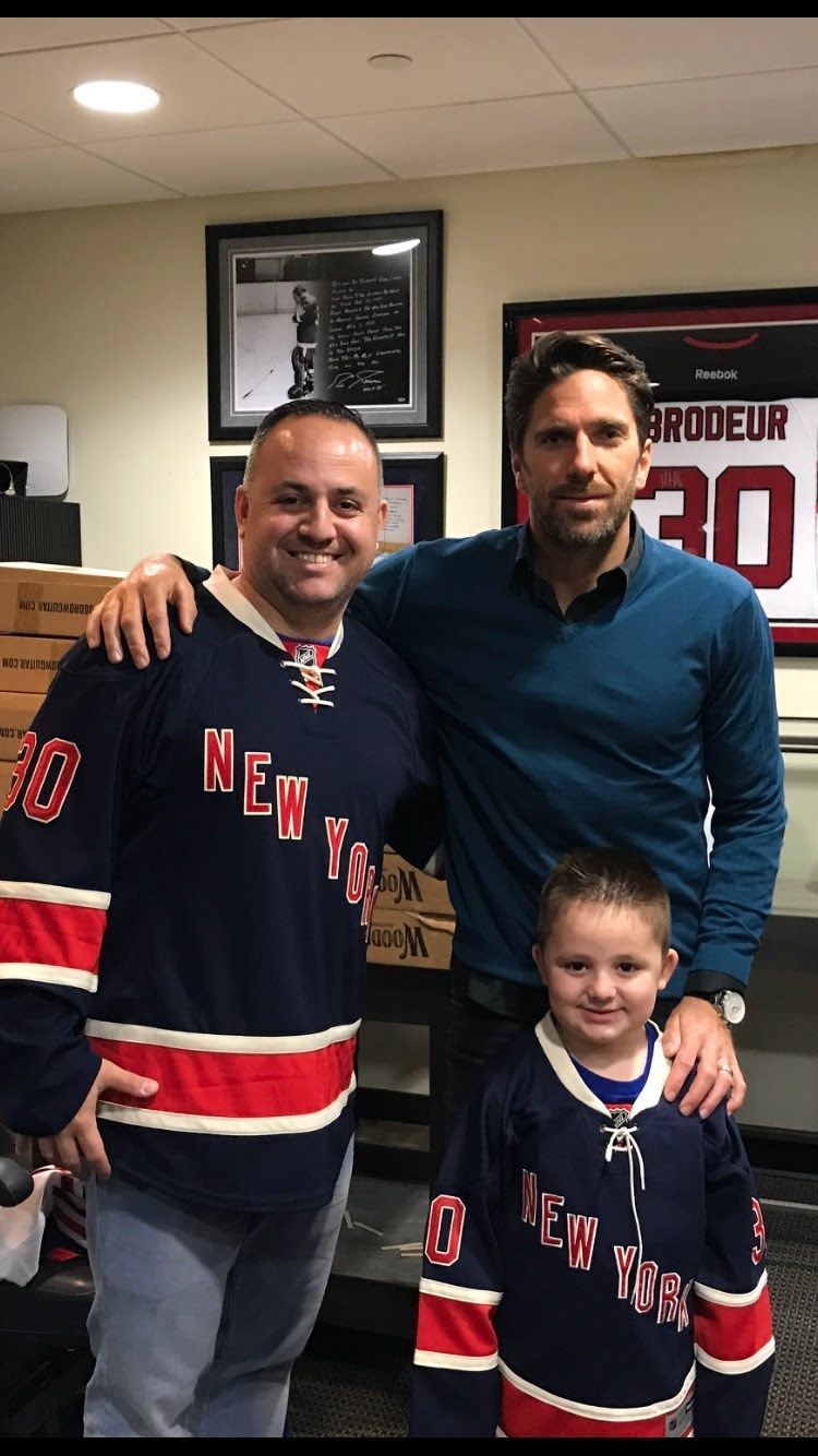 Man and child in Rangers jerseys with a man. Background includes framed items and a hockey jersey.