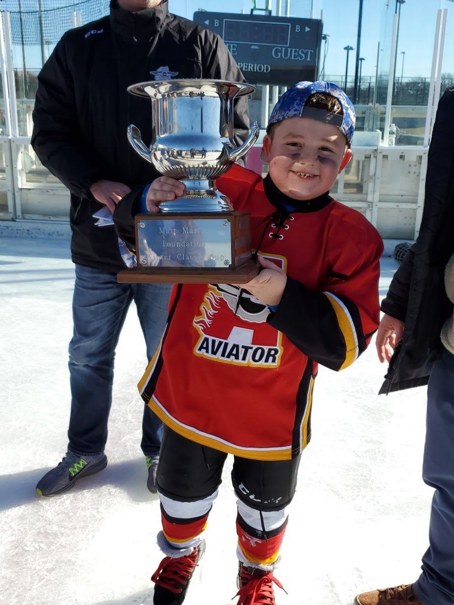 Young hockey player in red jersey holding a trophy, smiling. Outdoors on ice.
