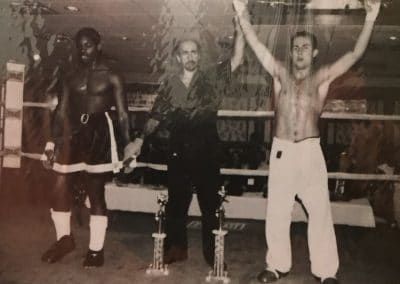 Boxers in a ring after a match, one arms raised in victory, flanked by an opponent and a referee, with trophies.