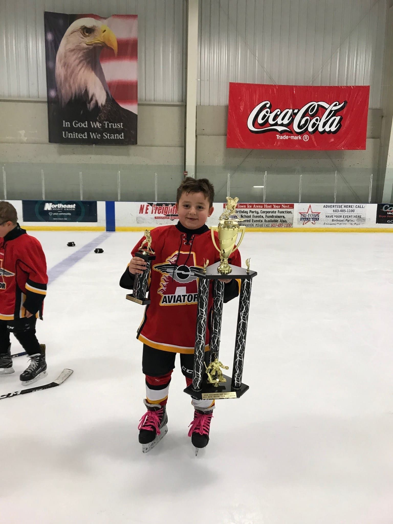 Boy in hockey gear on ice, holding trophies. He smiles, wearing red jersey and pink skate covers, inside an ice rink.