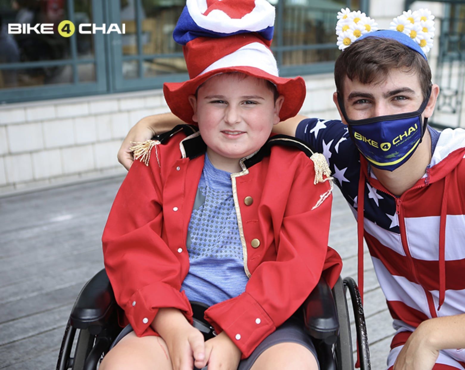 Boy in wheelchair, red outfit, smiles. Man with American flag shirt, smiles, arms around him. Outdoor setting.