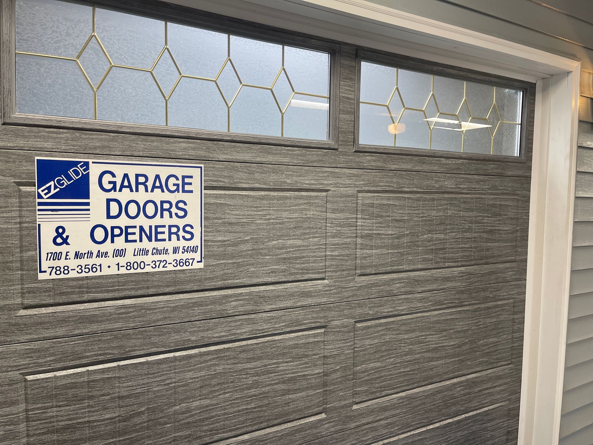 Gray garage door with a “Garage Doors & Openers” sign and decorative glass panels above.