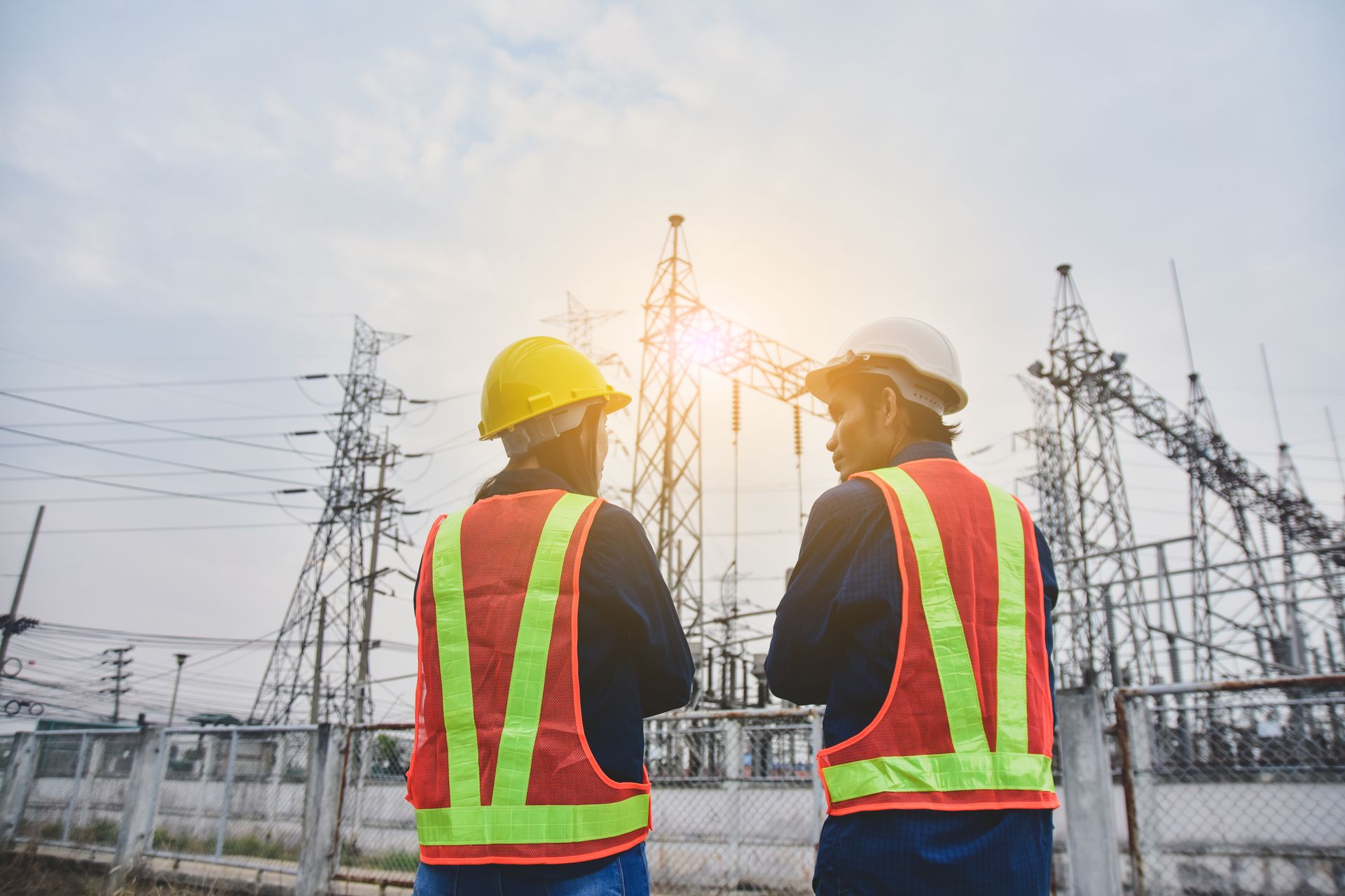 Two construction workers are standing next to each other in front of a power plant.
