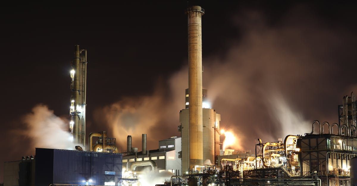 Night view of an industrial complex with illuminated structures, smoke, and a tall smokestack.