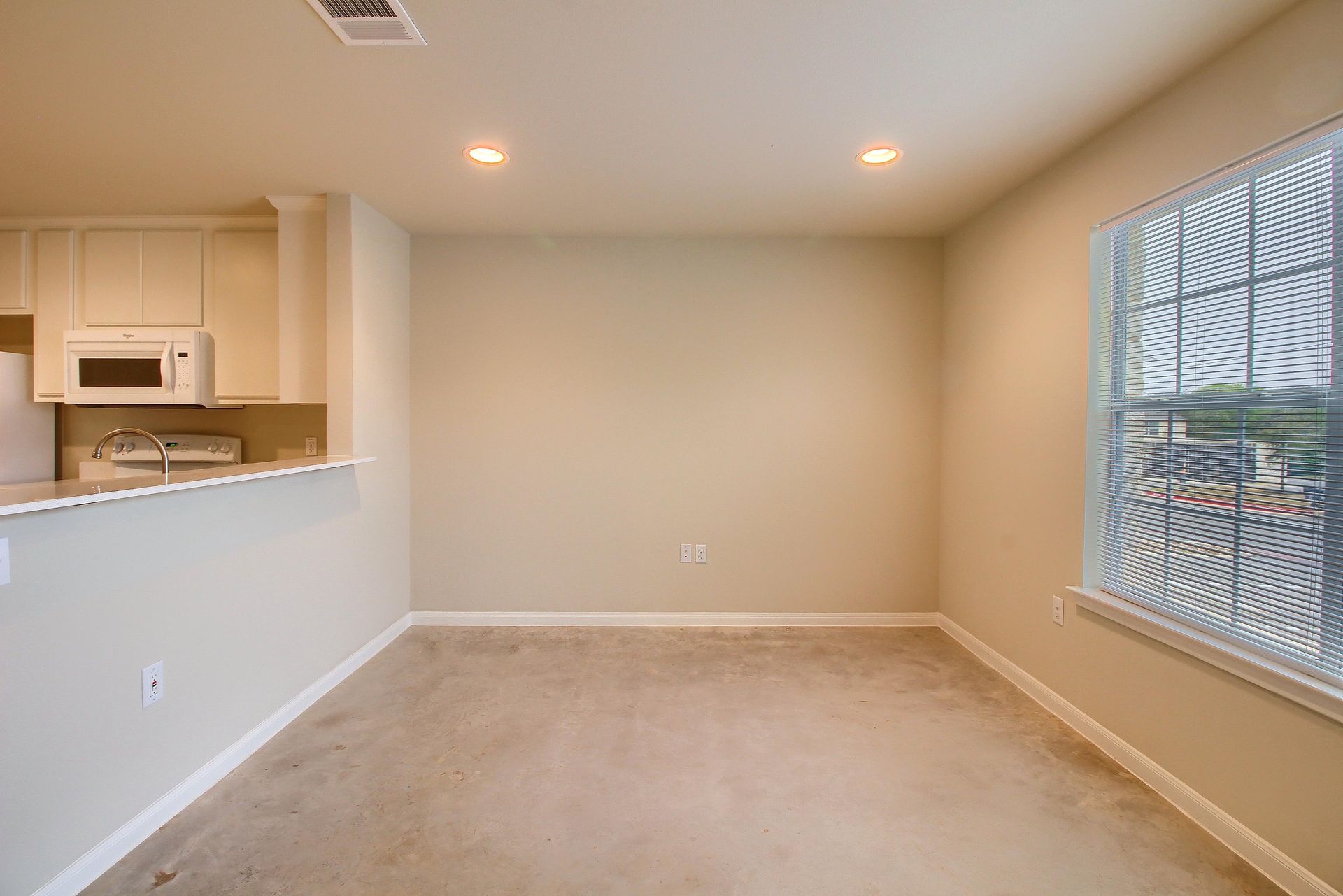 An empty living room with a window and a kitchen in the background.