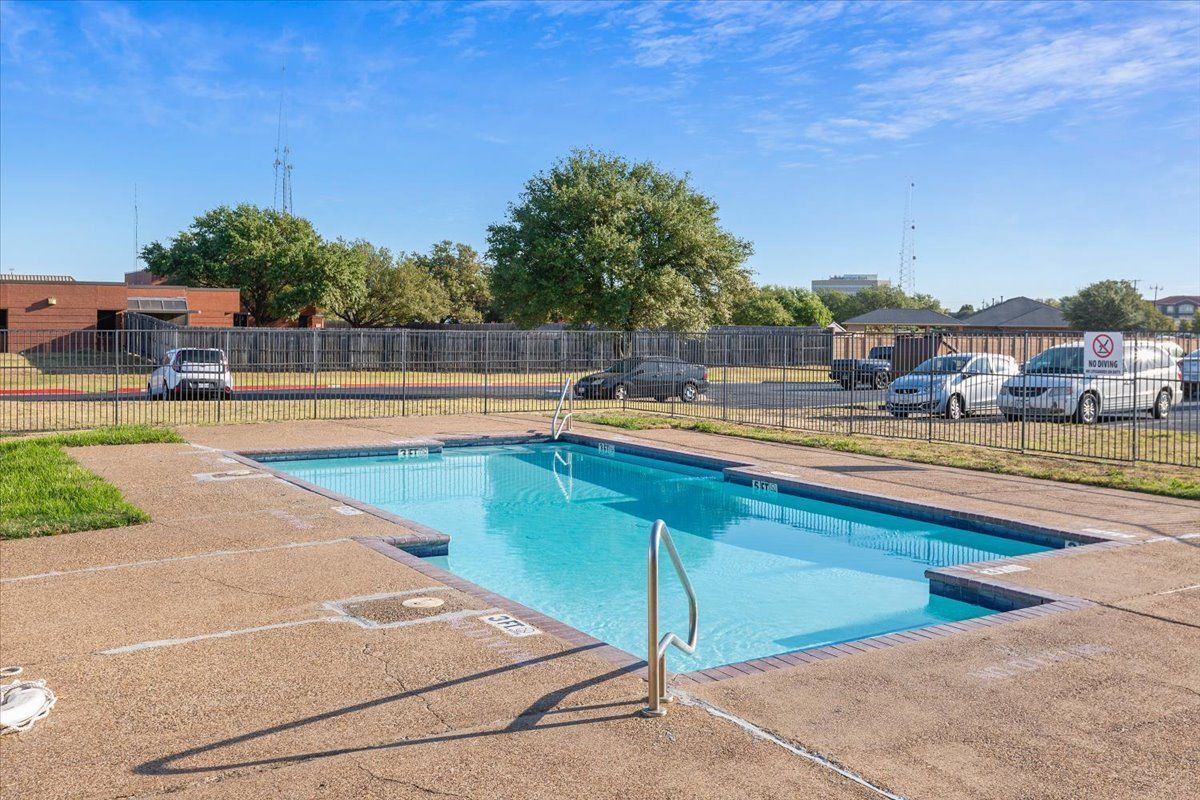 A large swimming pool is surrounded by a fence and cars parked in front of it.