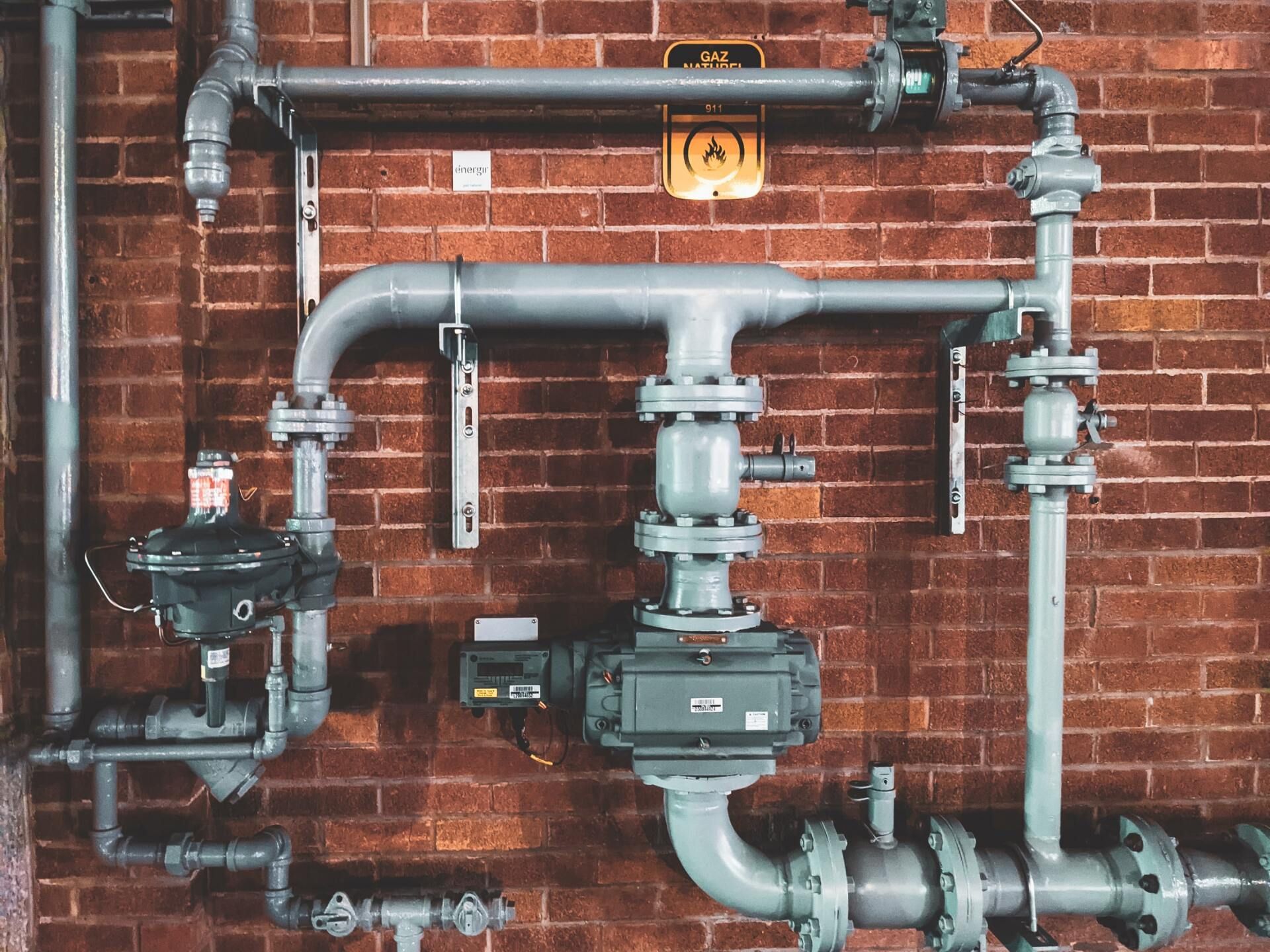 Grey industrial pipes and valves against a red brick wall.