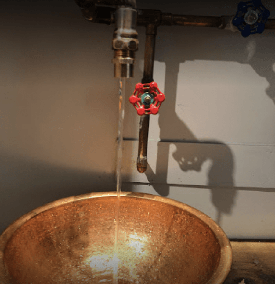 A copper sink with water running from a faucet with a red handle.