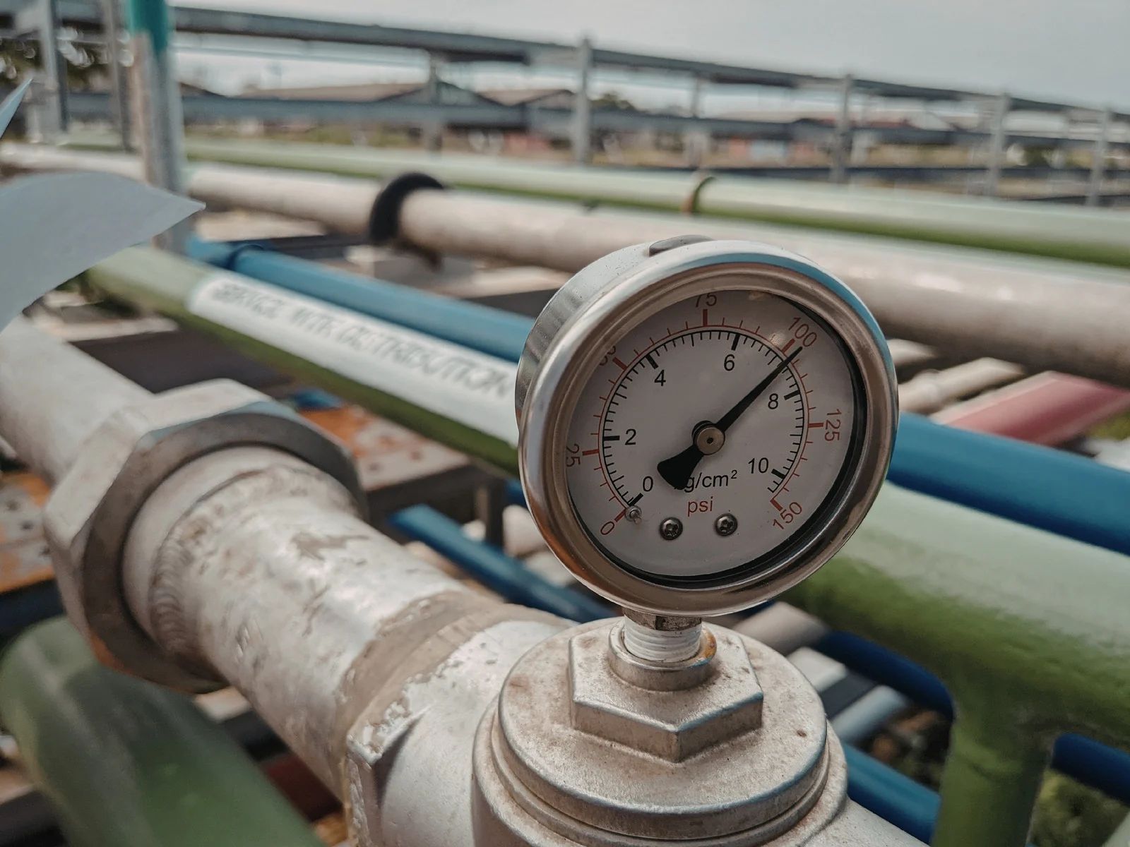 Pressure gauge on industrial pipes, reading approximately 5. Green and blue pipes in the background.