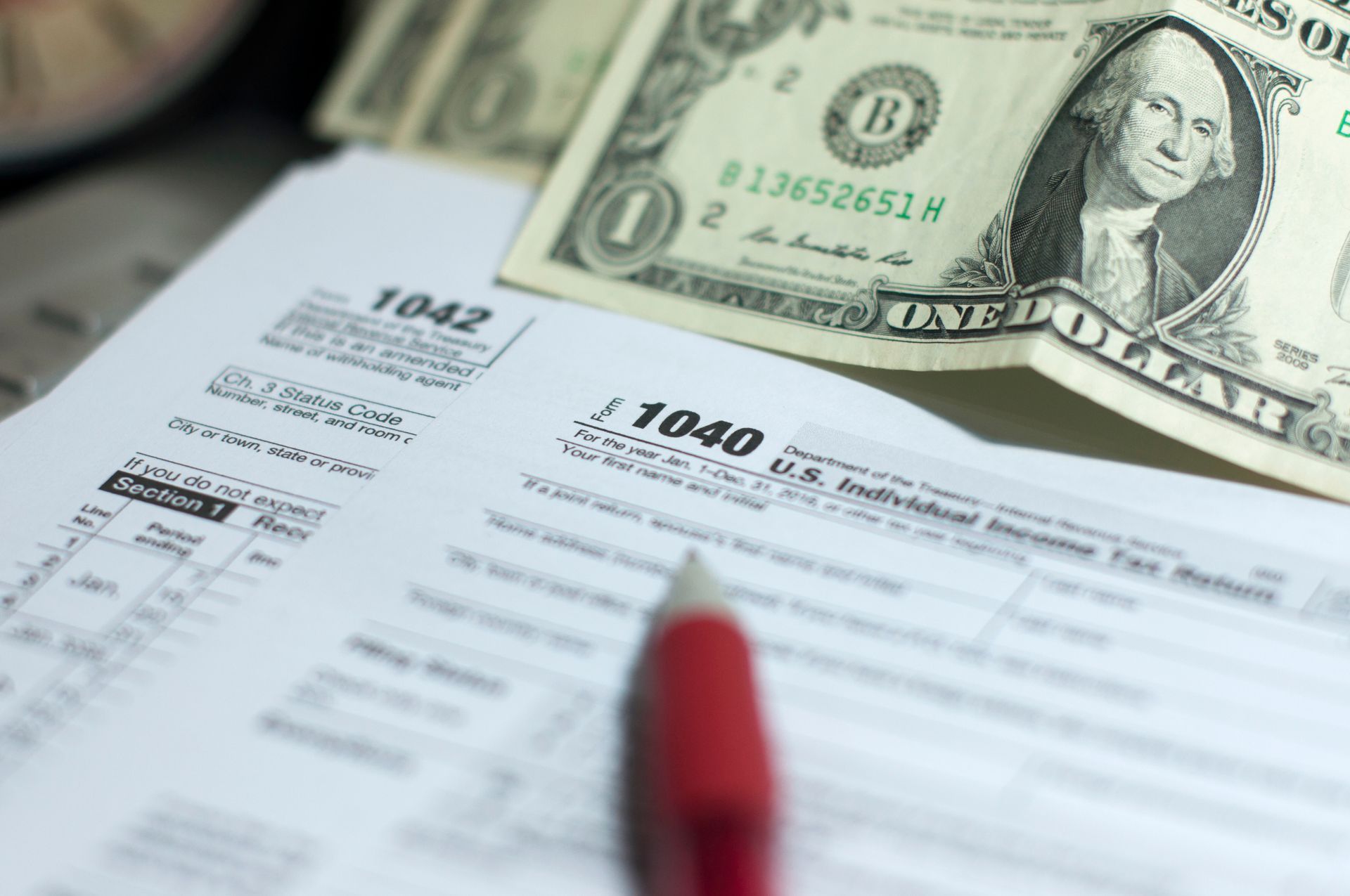 Tax forms (1040, 1042) with a dollar bill and a pen.