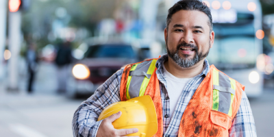 A construction worker is holding a hard hat and smiling.