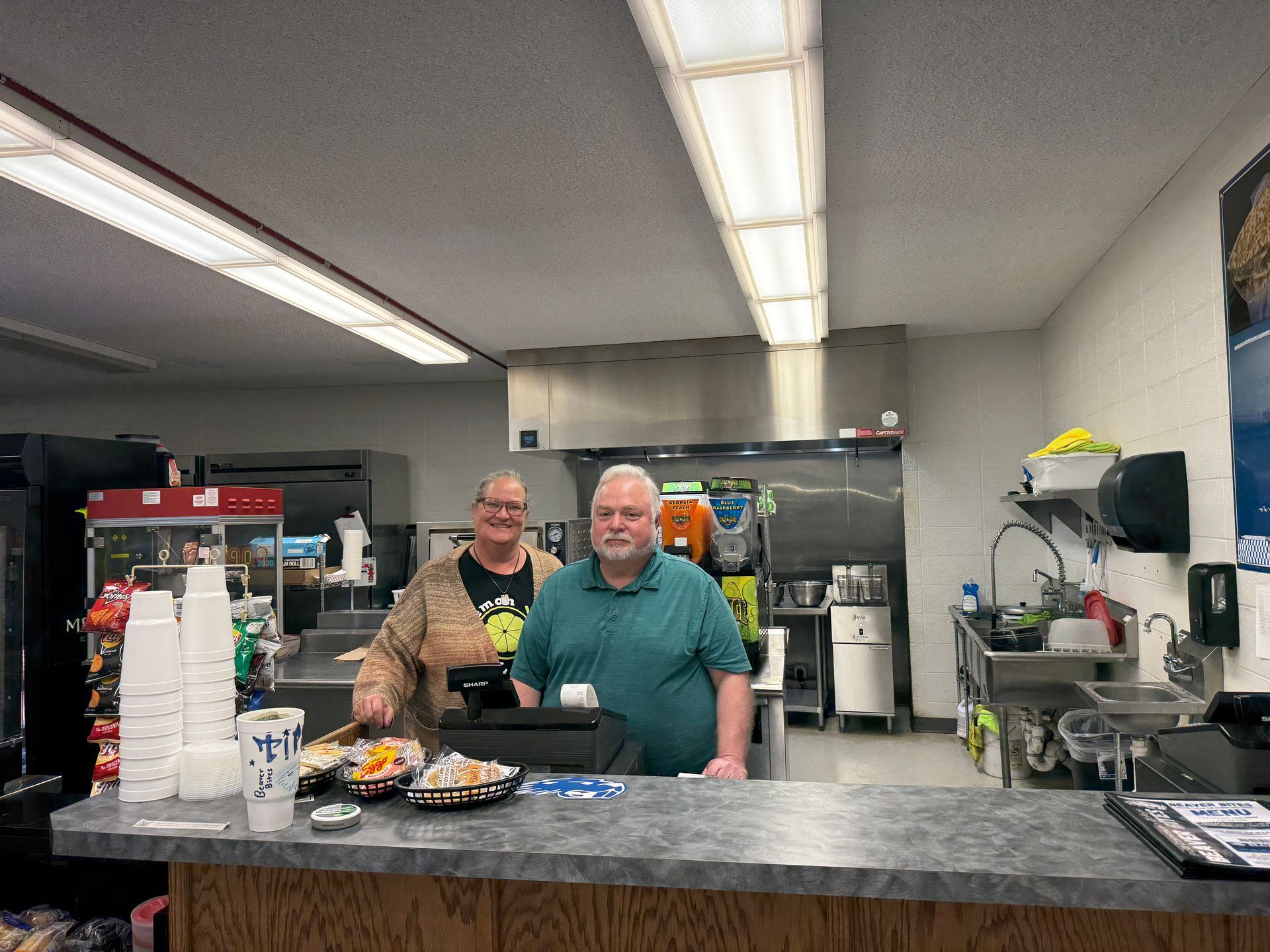Two people stand behind a counter in a small kitchen. Food and drink items are visible.