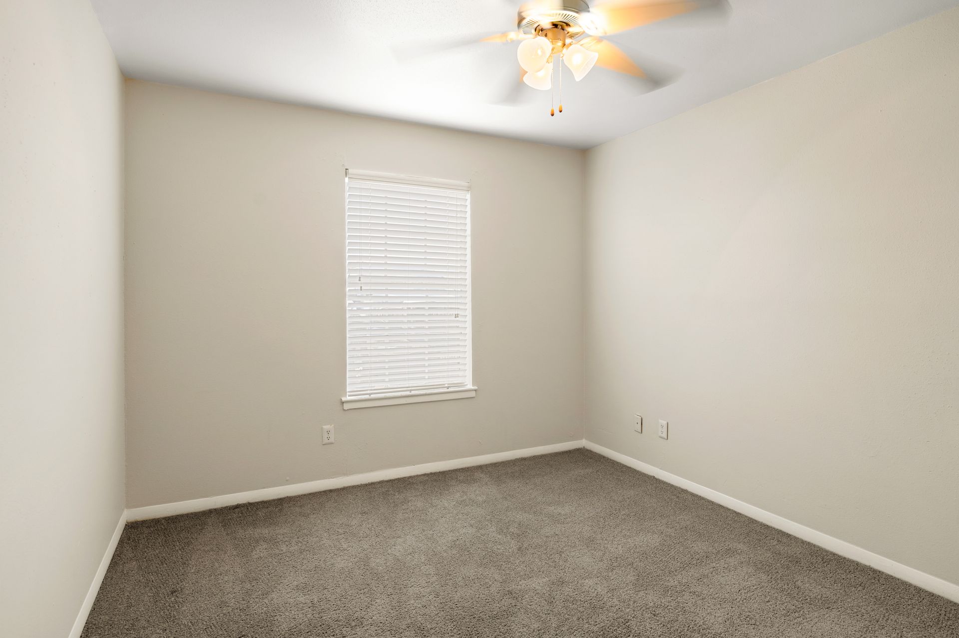 Empty bedroom with beige walls, carpet, and window blinds. Ceiling fan present.