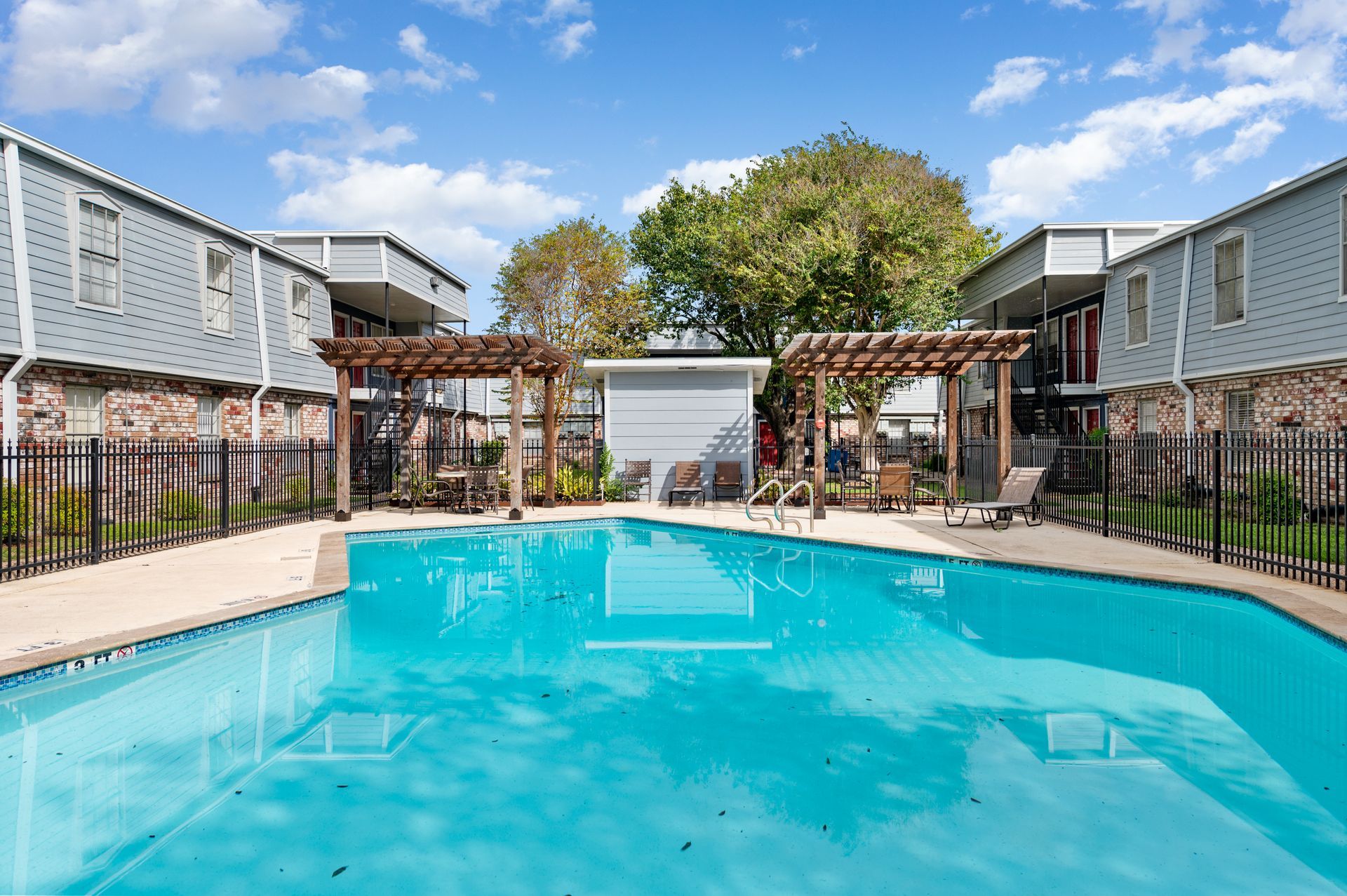 Swimming pool surrounded by two-story apartment buildings and a wooden pergola, under a blue sky with clouds.