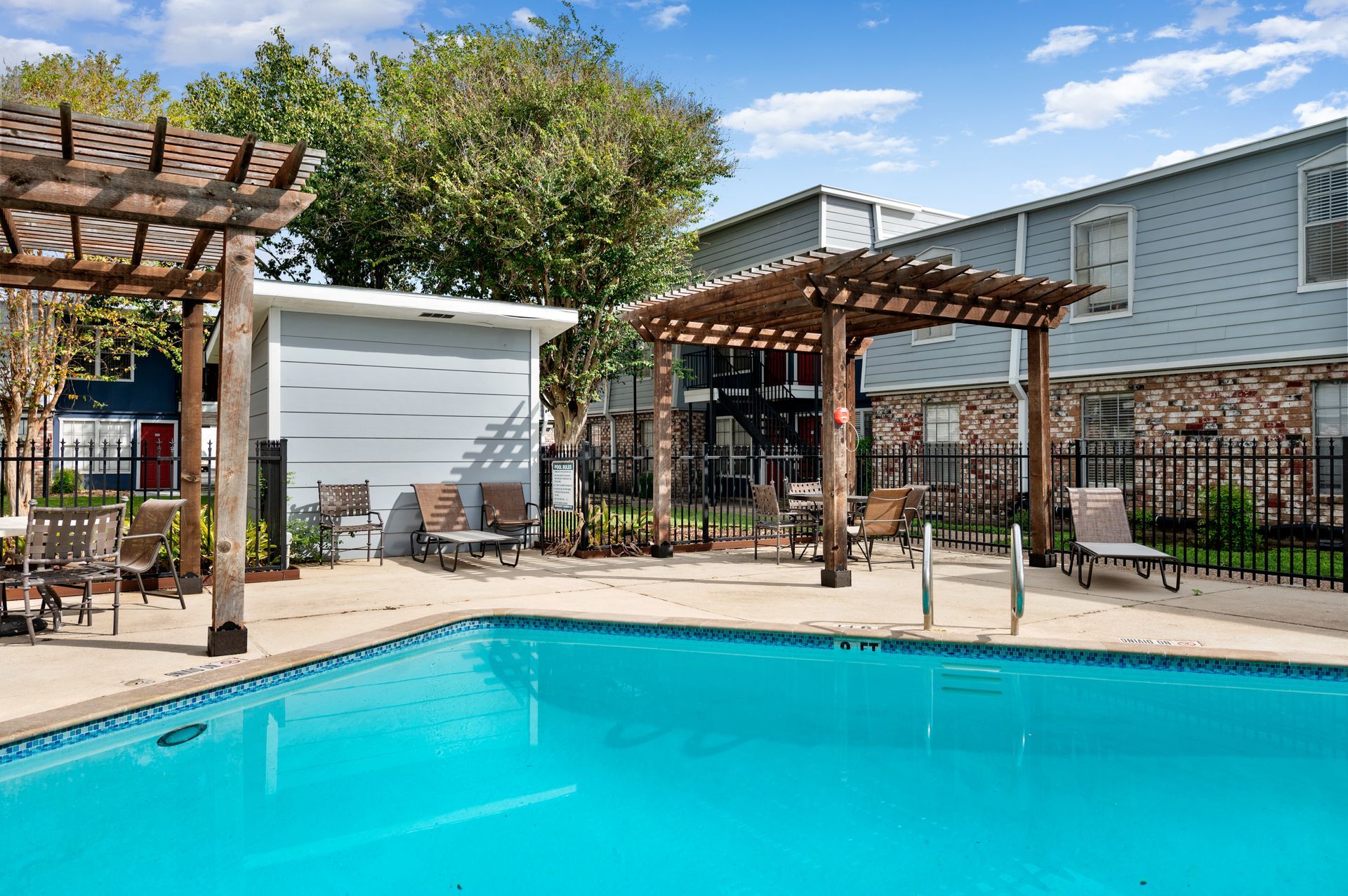 Pool area with pergola, lounge chairs, and two-story apartment building under a blue sky.