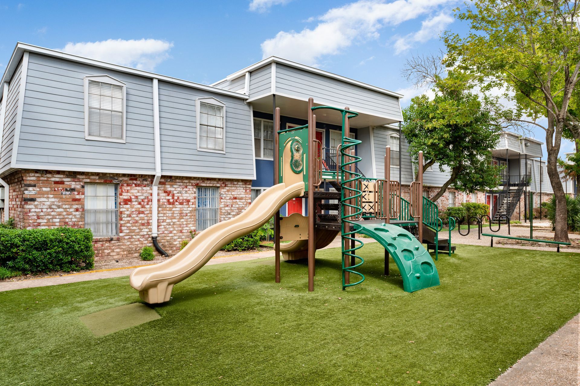 Playground in front of two-story apartment building with beige slide, green climbing elements, and artificial turf.