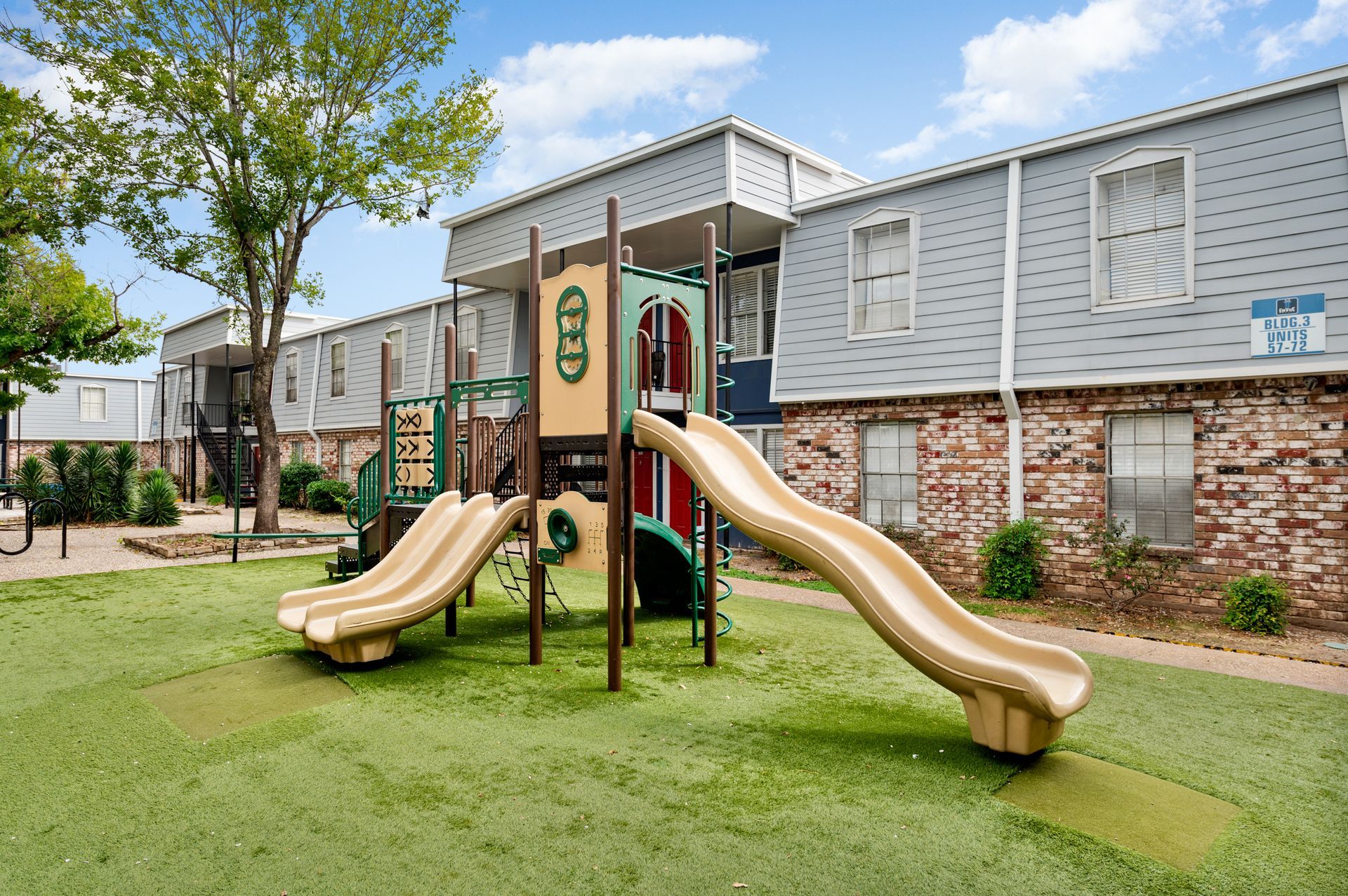 Playground in front of two-story apartment building with tan slides, green turf, and trees under a blue sky.