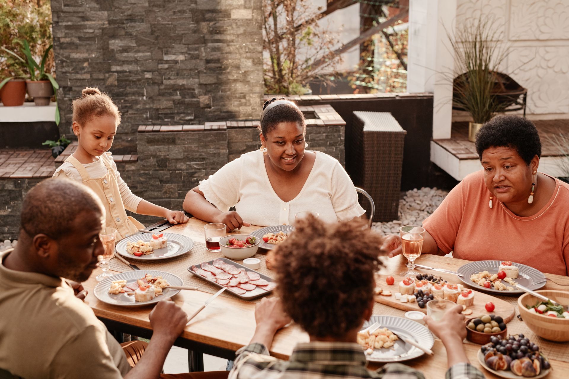 Family gathered around a wooden table, enjoying a meal outdoors. Plates of food and fruit are visible.