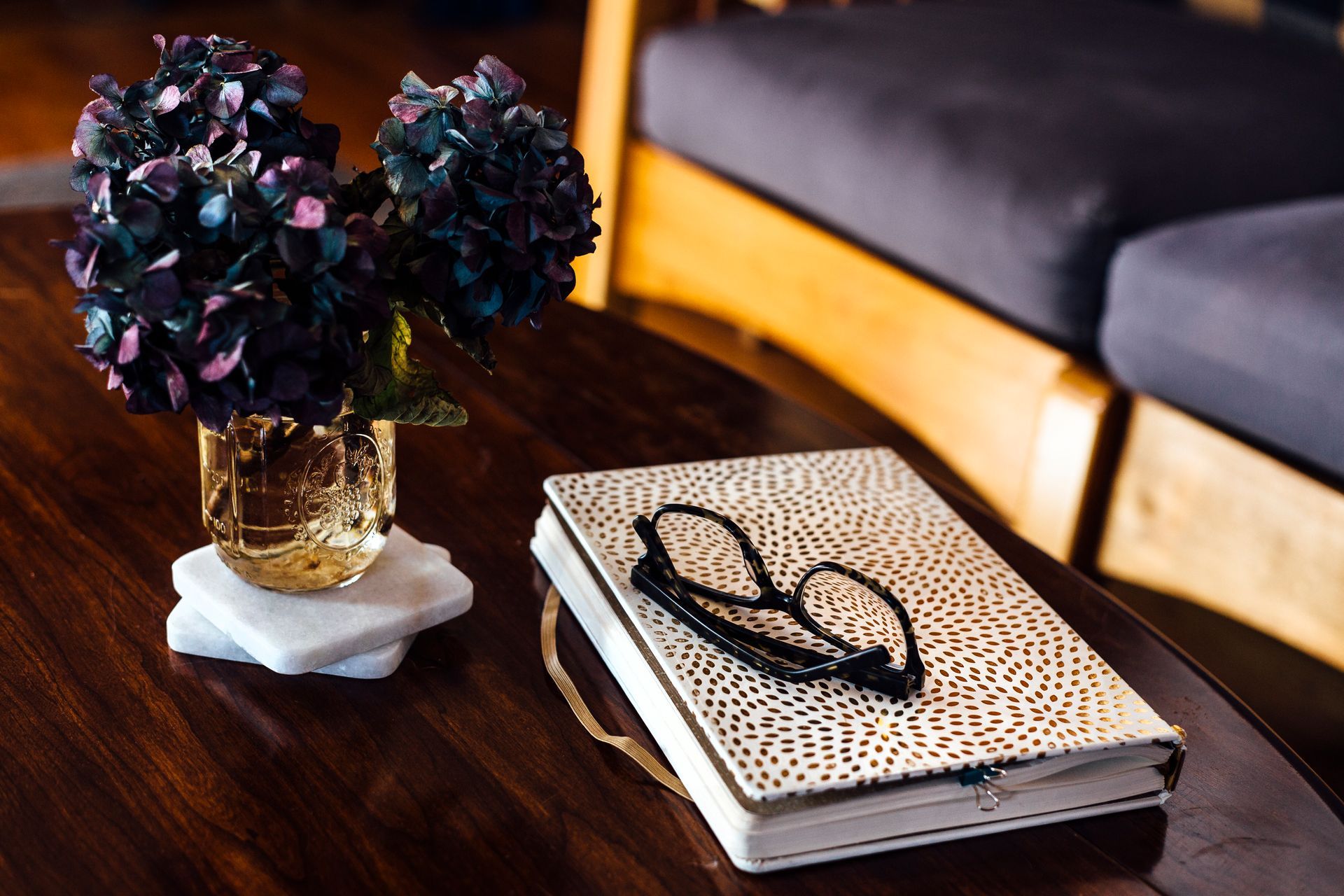 Dark wood coffee table with flowers, a journal, and glasses; a couch in the background.