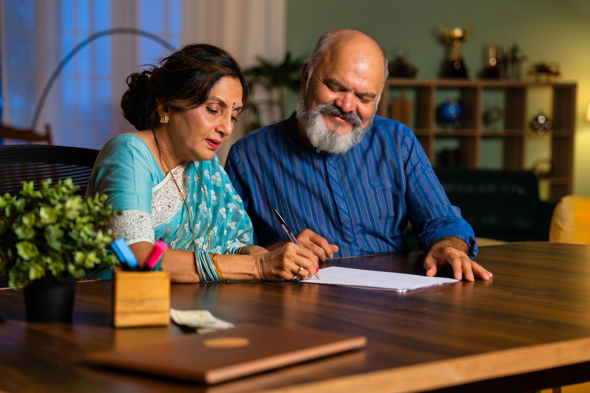 Couple signing document at a wooden table. The woman wears a blue sari, the man a blue shirt.