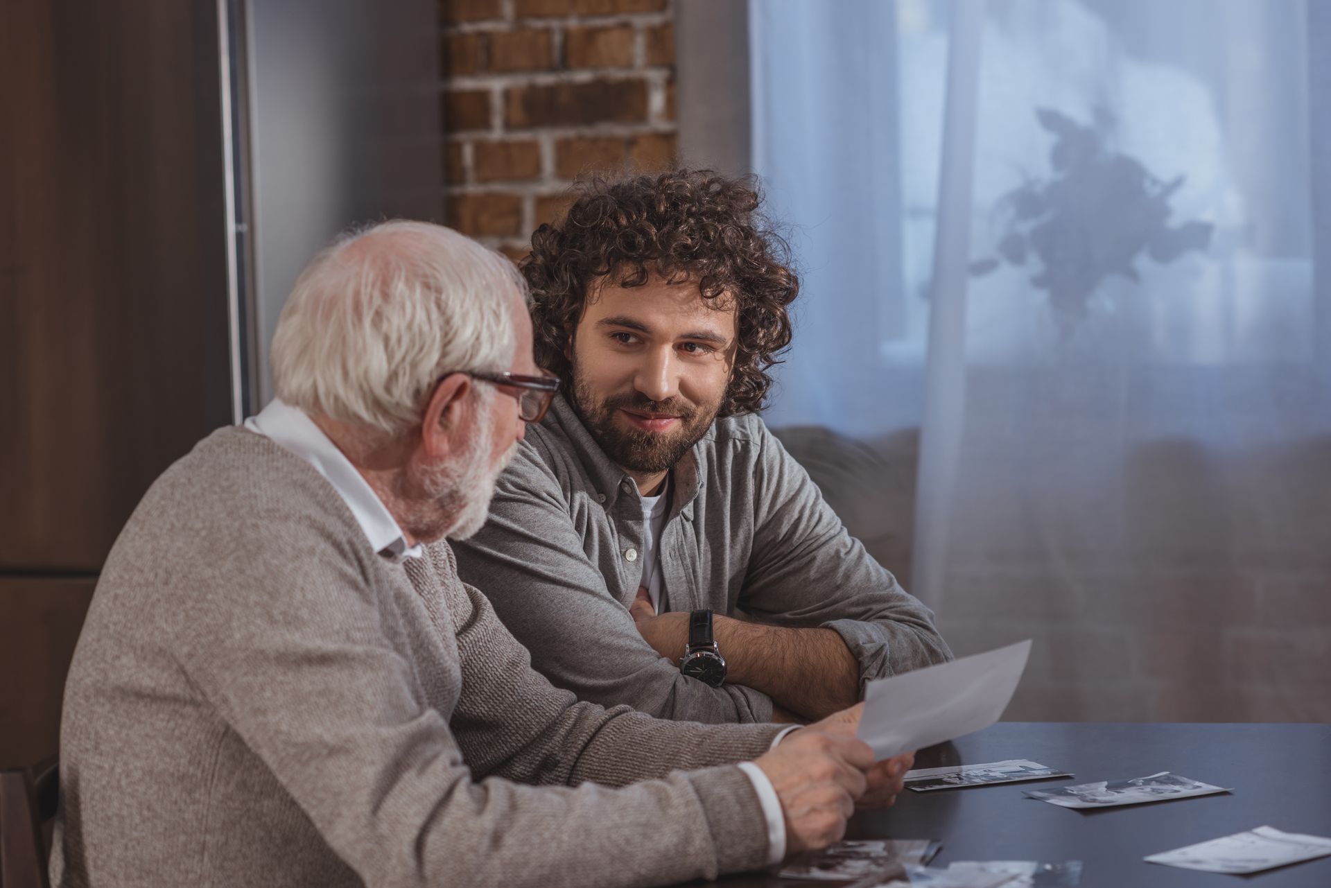Older man with glasses showing documents to a younger man with curly hair; they are seated at a table.Icon
